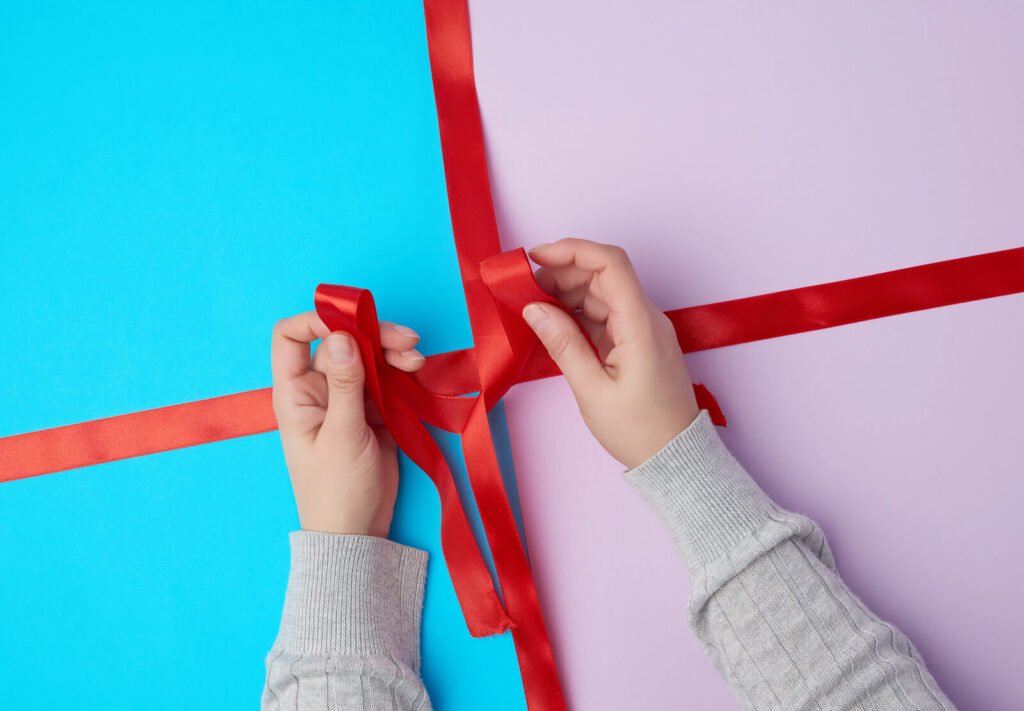 Hands tie a bow with a satin red ribbon on a gift box.